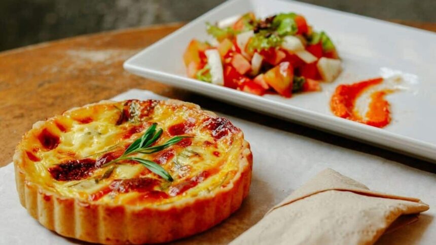 Quiche individual dourada com ramo de alecrim, acompanhada de salada e molho em um prato branco.