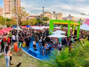 Multidão em festival ao ar livre com barracas de comida, palco, balão rosa 'Belo Horizonte' e prédios.