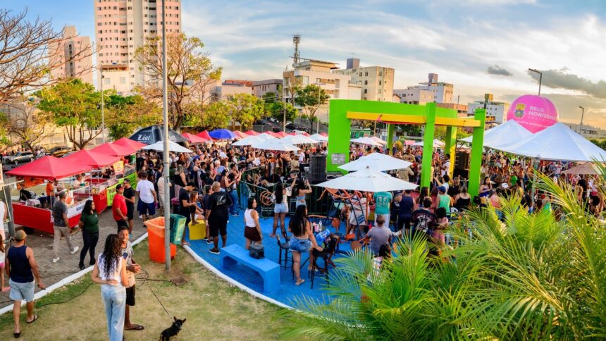 Multidão em festival ao ar livre com barracas de comida, palco, balão rosa 'Belo Horizonte' e prédios.