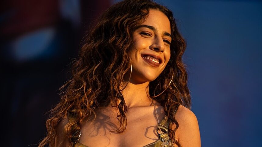 Close-up de mulher sorrindo, com cabelos cacheados e brincos de argola, em palco iluminado.