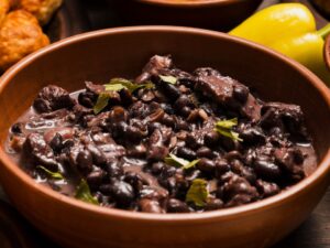 Close-up de tigela rústica com feijão preto, carne e folhas verdes, em mesa de madeira.