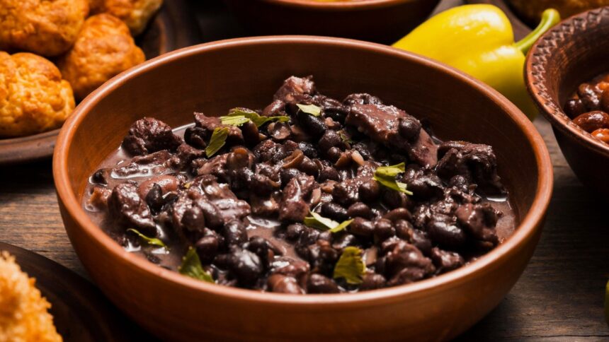 Close-up de tigela rústica com feijão preto, carne e folhas verdes, em mesa de madeira.