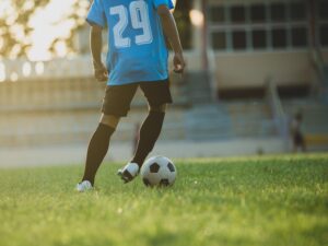 Jogador com camisa azul 29, shorts e meias pretas, tocando uma bola de futebol no gramado