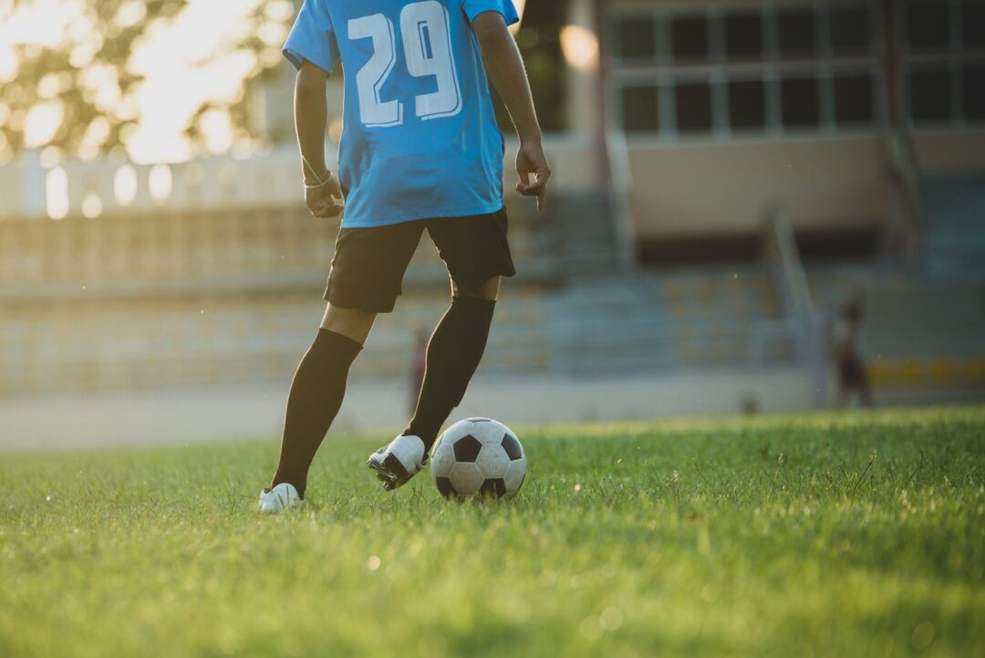 Jogador com camisa azul 29, shorts e meias pretas, tocando uma bola de futebol no gramado