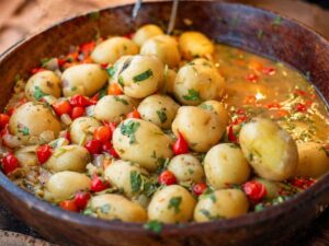 Cumbuca rústica de madeira com batatas pequenas em caldo, pimentas vermelhas e ervas frescas.
