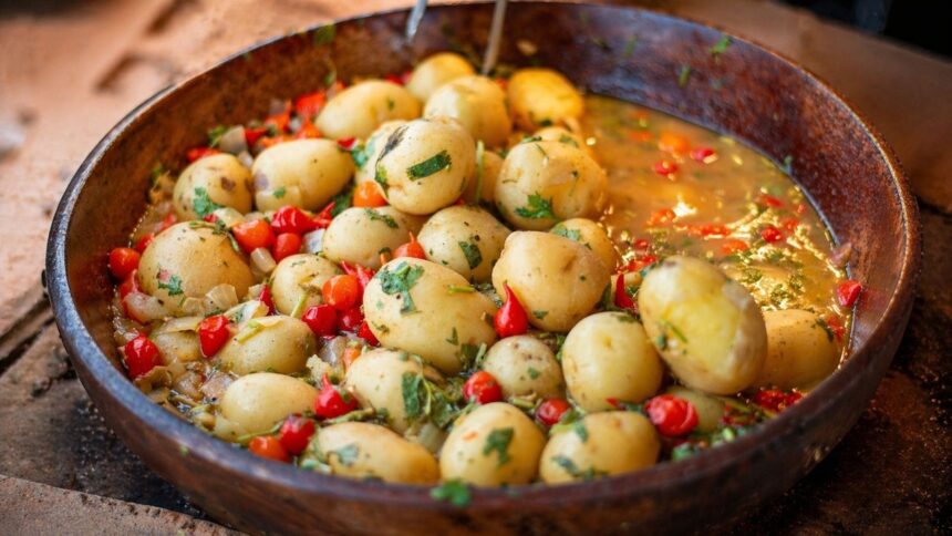 Cumbuca rústica de madeira com batatas pequenas em caldo, pimentas vermelhas e ervas frescas.
