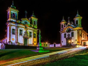 Duas igrejas barrocas iluminadas à noite e monumento central em praça de cidade histórica.