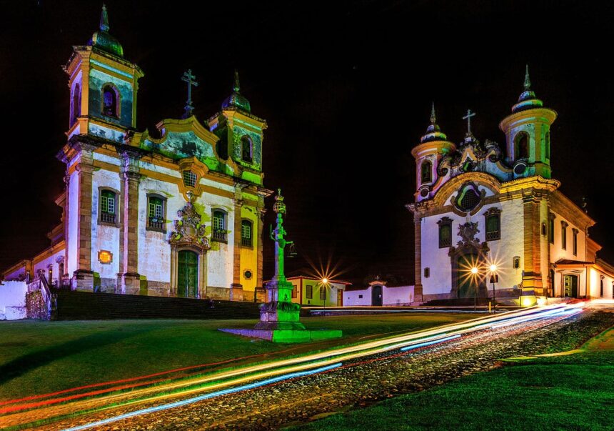 Duas igrejas barrocas iluminadas à noite e monumento central em praça de cidade histórica.