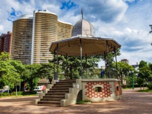 Coreto de metal e tijolo em praça com prédios altos e árvores verdes ao redor.