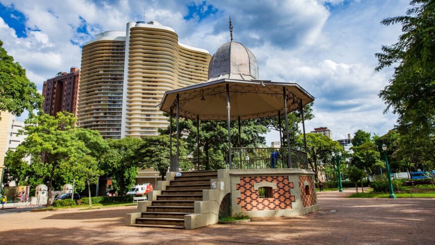 Coreto de metal e tijolo em praça com prédios altos e árvores verdes ao redor.