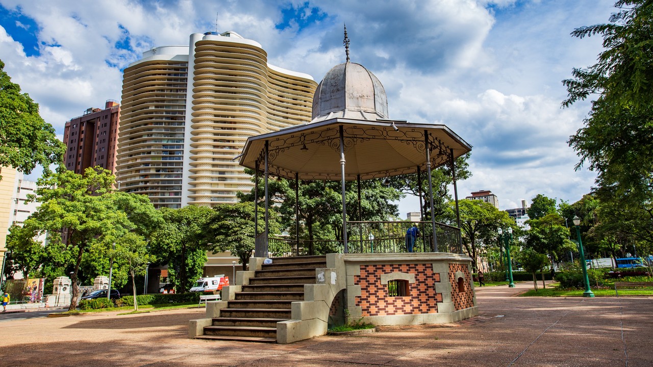 Coreto de metal e tijolo em praça com prédios altos e árvores verdes ao redor.