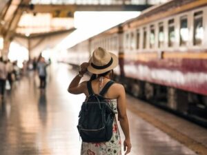 Mulher de chapéu e mochila em plataforma de trem, de costas, olhando para vagão.