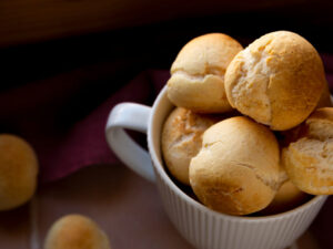 Close-up de pães de queijo dourados e redondos em uma caneca branca, com alguns espalhados.