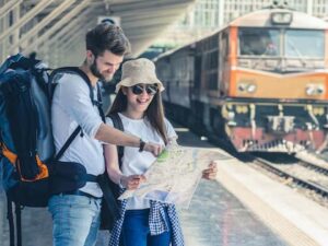 Casal viajante com mochilas olha mapa em estação de trem com locomotiva ao fundo.