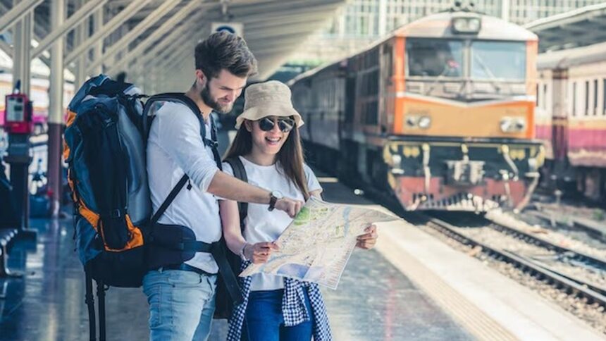 Casal viajante com mochilas olha mapa em estação de trem com locomotiva ao fundo.