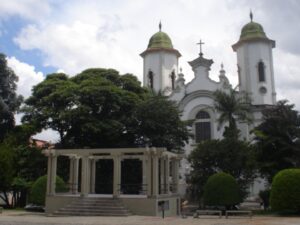 Igreja branca com torres e cúpulas verdes, coreto e árvores em praça com céu nublado.