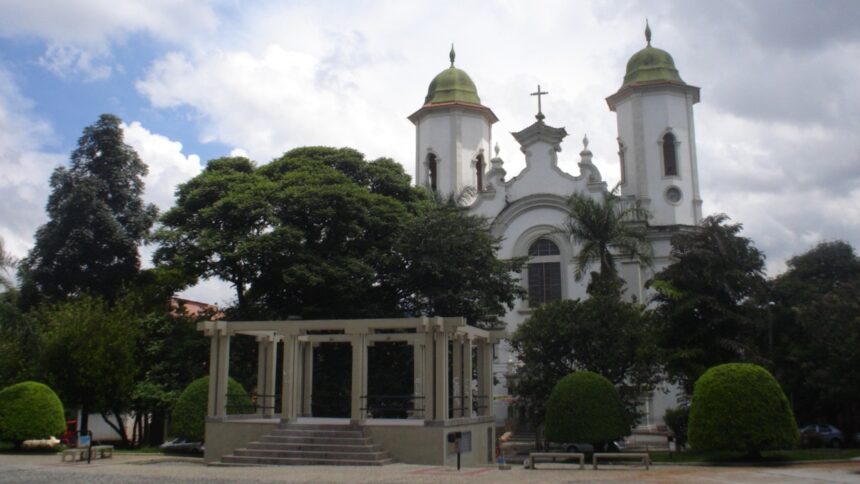 Igreja branca com torres e cúpulas verdes, coreto e árvores em praça com céu nublado.