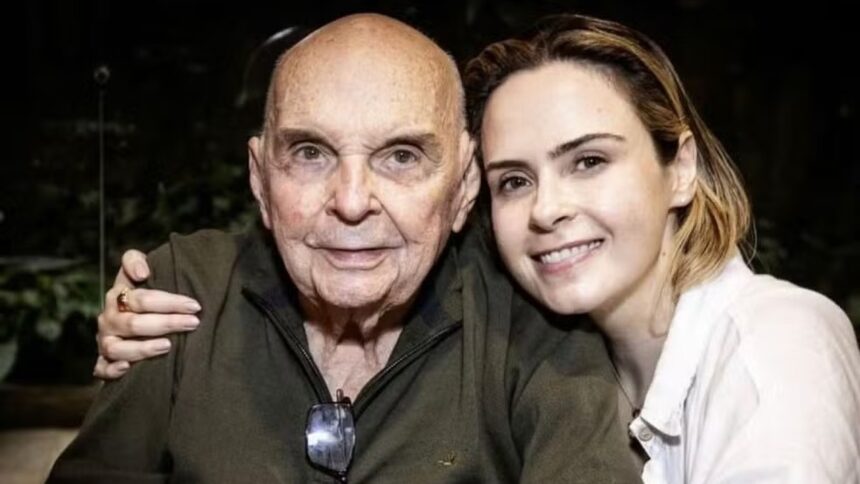 Homem idoso e mulher jovem, ambos sorrindo, abraçados. Mulher usa anel no dedo.