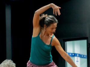 Mulher com top verde-azulado e cabelo preso dançando com braços levantados em fundo escuro.