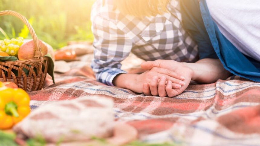 Mãos de casal entrelaçadas em toalha xadrez com cesta de piquenique e frutas.