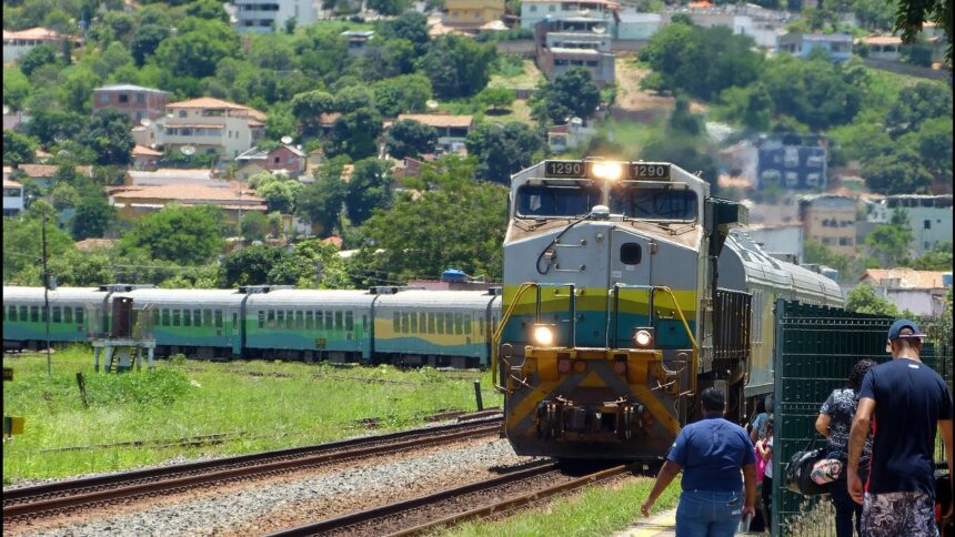 Locomotiva 1290 cinza e amarela puxa vagões verdes em trilhos, com pessoas e casas ao fundo.