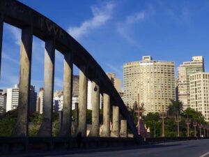 Ponte de concreto com pilares em arco, prédios altos em cidade urbana, sob céu azul.