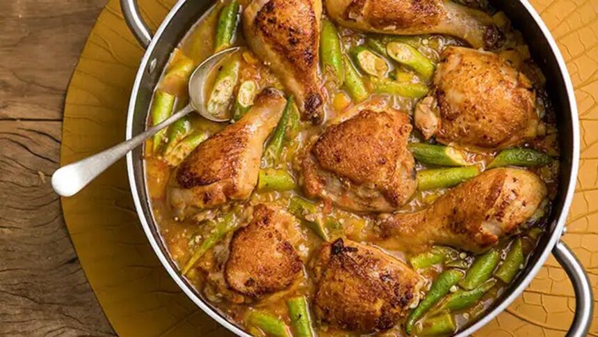 Frango com quiabo em panela, com colher, sobre base de madeira e dourada.