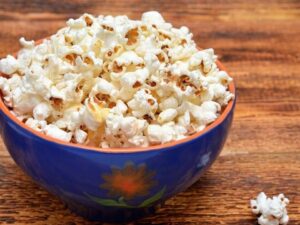 Tigela azul cheia de pipoca sobre mesa de madeira, com grãos espalhados.