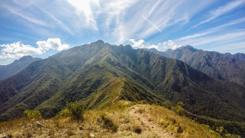 Caminho de terra em cume de montanha arborizada sob céu azul com nuvens.