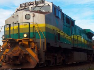 Locomotiva de trem 1225, verde, amarela e cinza, nos trilhos sob céu azul.