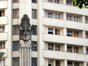 Escultura de um rosto indígena em destaque na fachada de um edifício de cor clara com janelas.