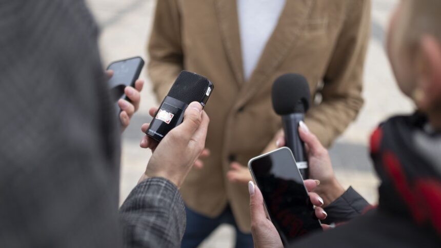 Mãos seguram microfones e smartphones registrando áudio e vídeo em uma entrevista.
