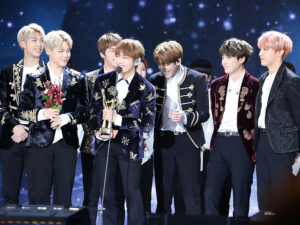 Sete membros do BTS no palco, sorrindo e celebrando com troféu e flores.