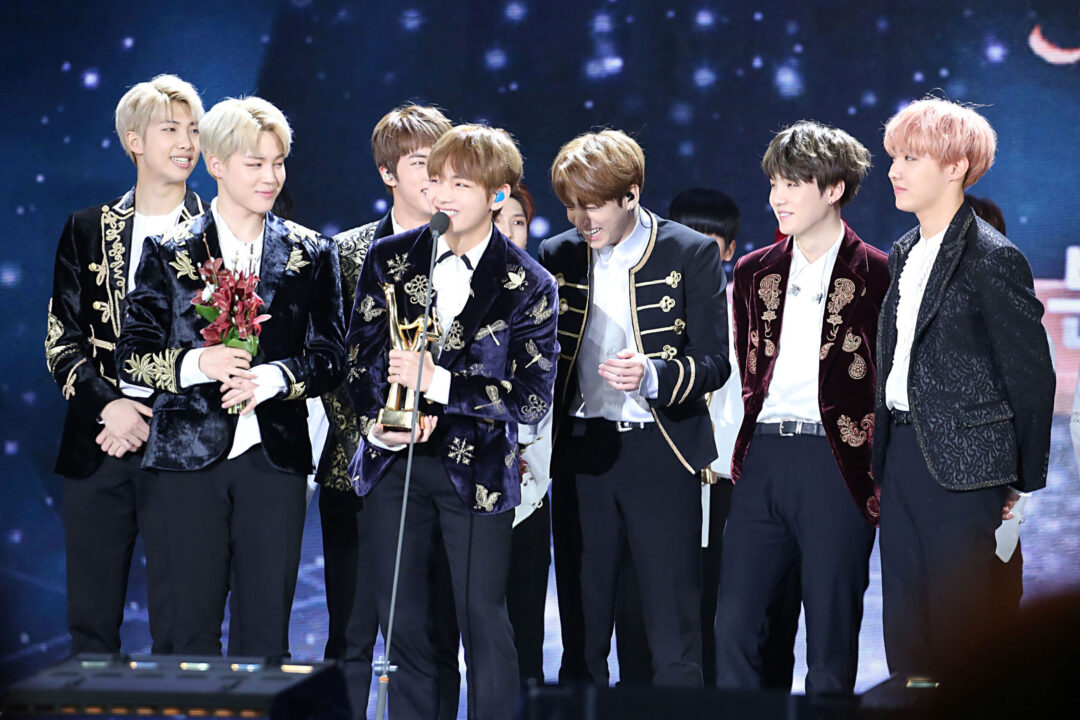 Sete membros do BTS no palco, sorrindo e celebrando com troféu e flores.