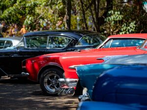 Fileira de carros antigos coloridos, vermelho e preto, estacionados ao ar livre com pessoas ao fundo