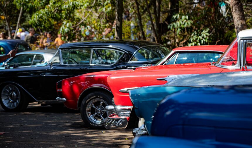 Fileira de carros antigos coloridos, vermelho e preto, estacionados ao ar livre com pessoas ao fundo