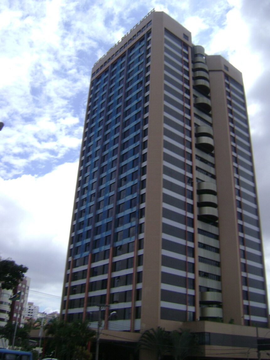 Prédio alto moderno com janelas escuras, faixas brancas e pilares marrons sob céu nublado.