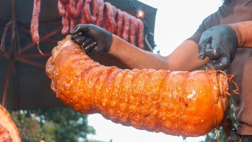 Homem com luvas pretas segura porco assado em rolo, com linguiças penduradas ao fundo.