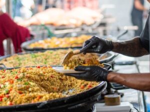 Pessoa de luvas pretas serve arroz de uma panela grande em evento.