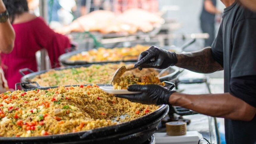 Pessoa de luvas pretas serve arroz de uma panela grande em evento.
