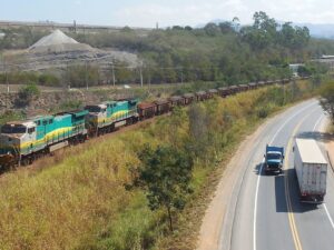 Trem de carga verde e amarelo em trilhos, paralelo a uma rodovia com dois caminhões.