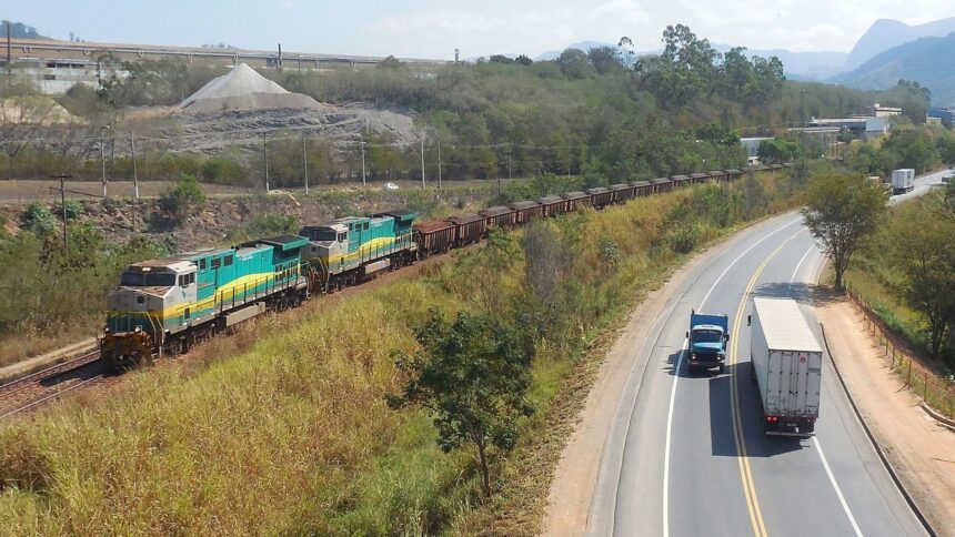 Trem de carga verde e amarelo em trilhos, paralelo a uma rodovia com dois caminhões.
