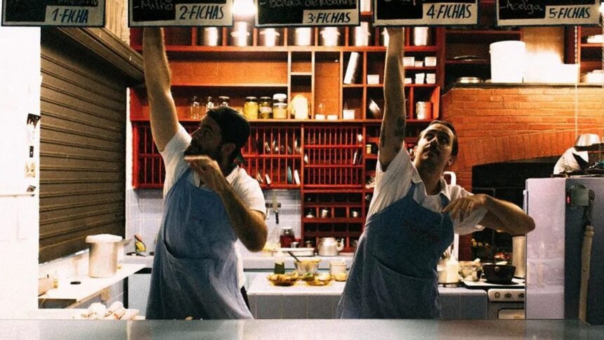 Dois cozinheiros com aventais seguram placas de menu 'FICHAS' e itens de comida em cozinha de mercado.