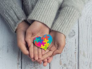 Mãos seguram um coração colorido em formato de quebra-cabeça, símbolo associado à conscientização sobre o autismo