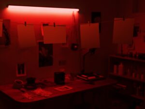 Darkroom banhado em luz vermelha, com papéis fotográficos pendurados e equipamentos sobre uma mesa
