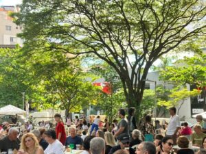 Pessoas em mesas ao ar livre sob árvores frondosas. Edifícios, tendas e uma placa "ESTACIONAM" ao fundo.