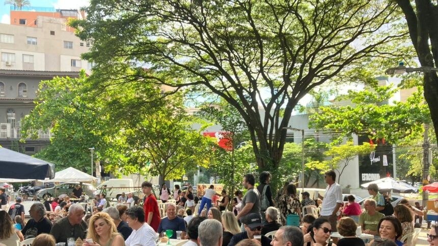 Pessoas em mesas ao ar livre sob árvores frondosas. Edifícios, tendas e uma placa "ESTACIONAM" ao fundo.