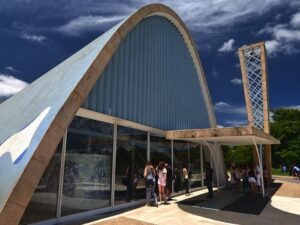 Exterior da Igreja da Pampulha, azul e curva, torre sineira e pessoas na entrada.