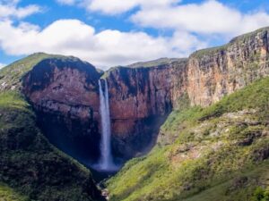 Alta cachoeira entre paredões rochosos verdejantes sob céu azul com nuvens brancas.
