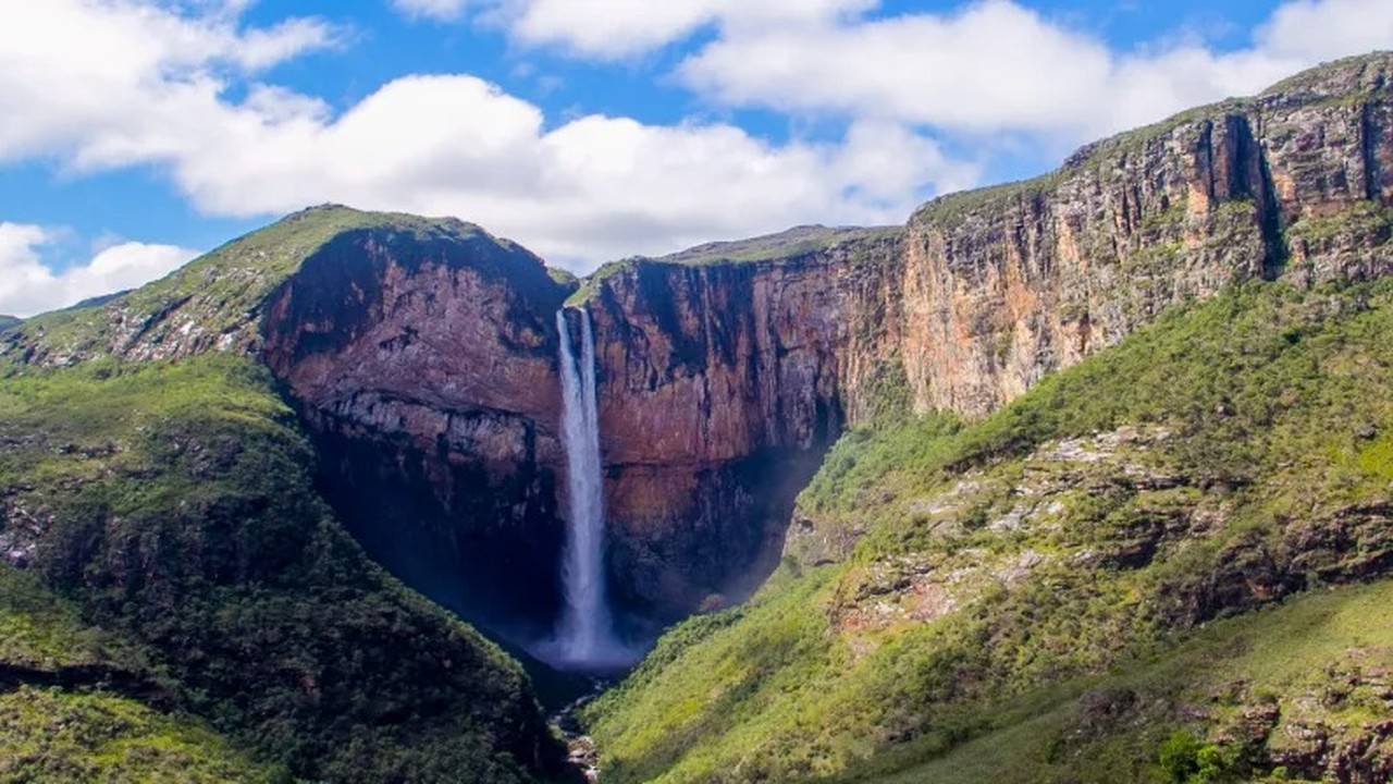 Alta cachoeira entre paredões rochosos verdejantes sob céu azul com nuvens brancas.